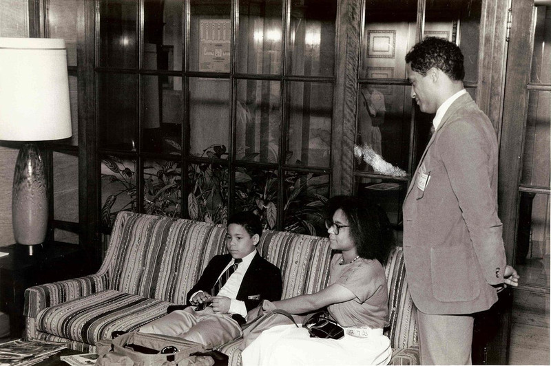 Black-and-white photograph of three people: two seated on a sofa and one standing beside them.
